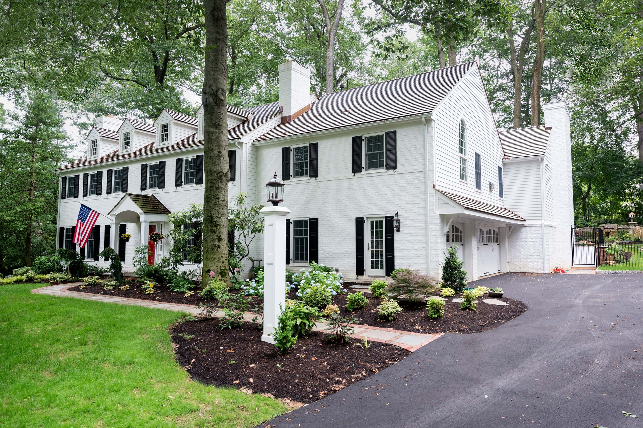 Charming white house with black shutters, American flag, lush landscaping, and driveway, surrounded by trees. Perfect suburban home aesthetic.