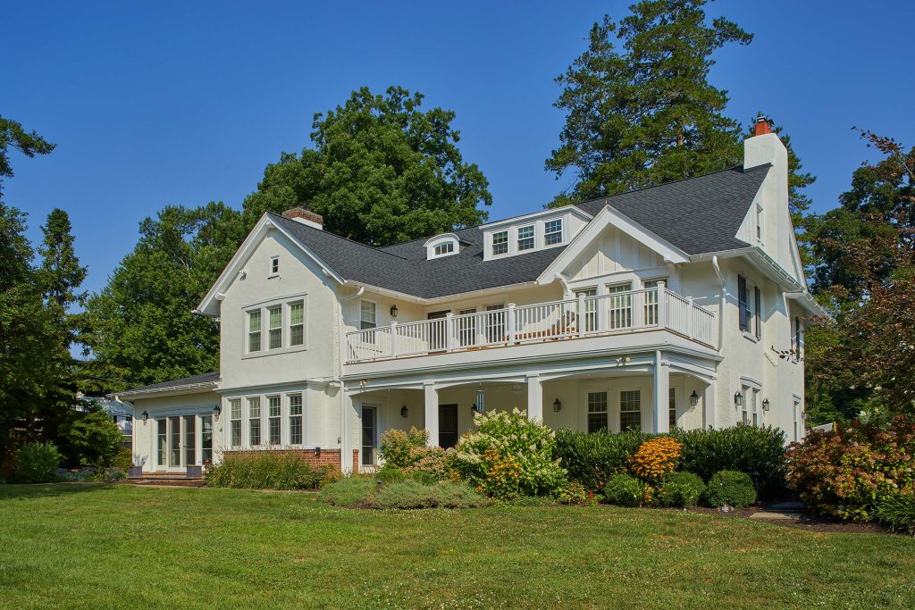 Yellow two-story house with wraparound porch, balcony, and dark roof, surrounded by green lawn, shrubs, and trees under a clear sky.