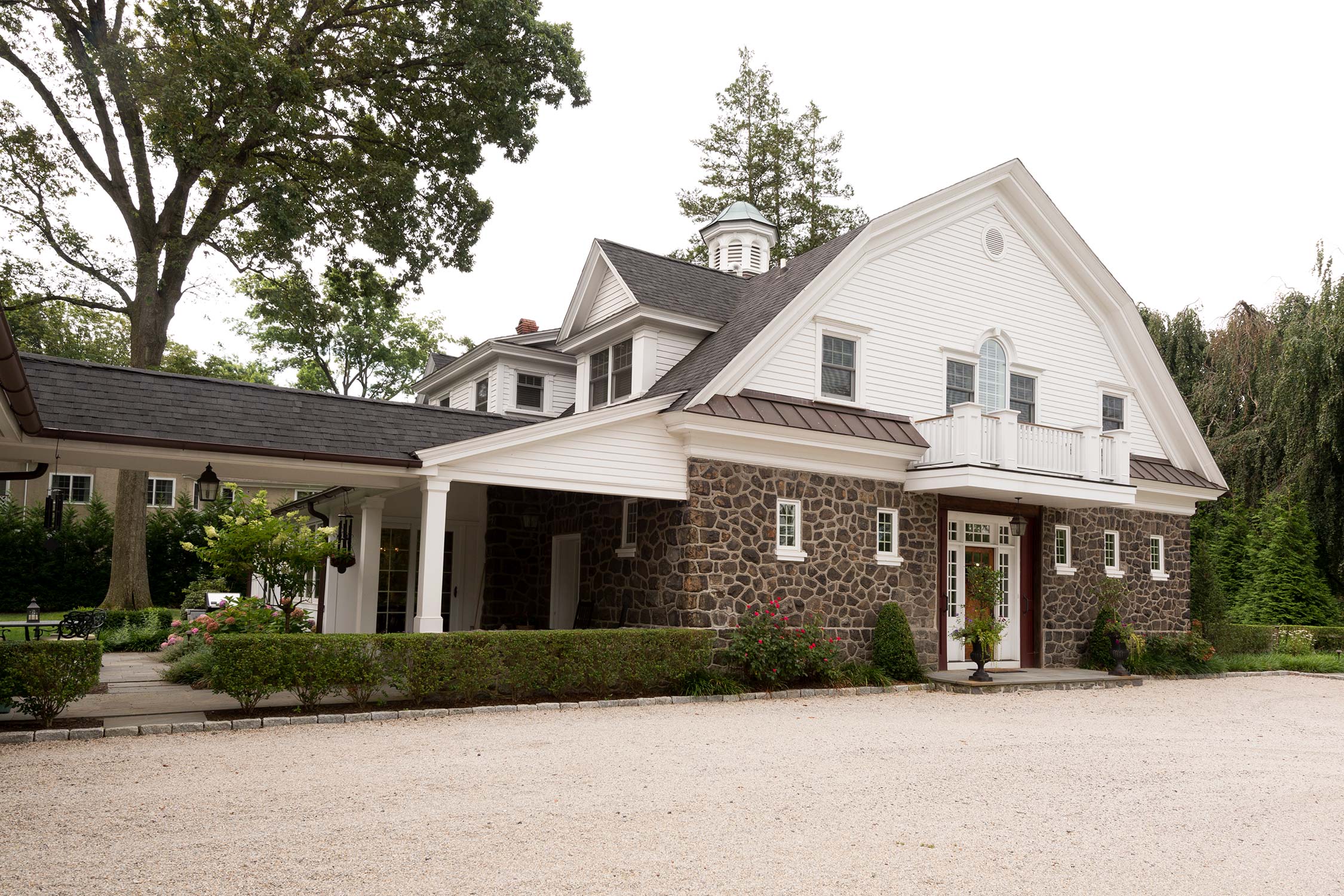 A stone and white barn-style house with a covered porch, balcony, and gravel driveway, surrounded by greenery and trees.