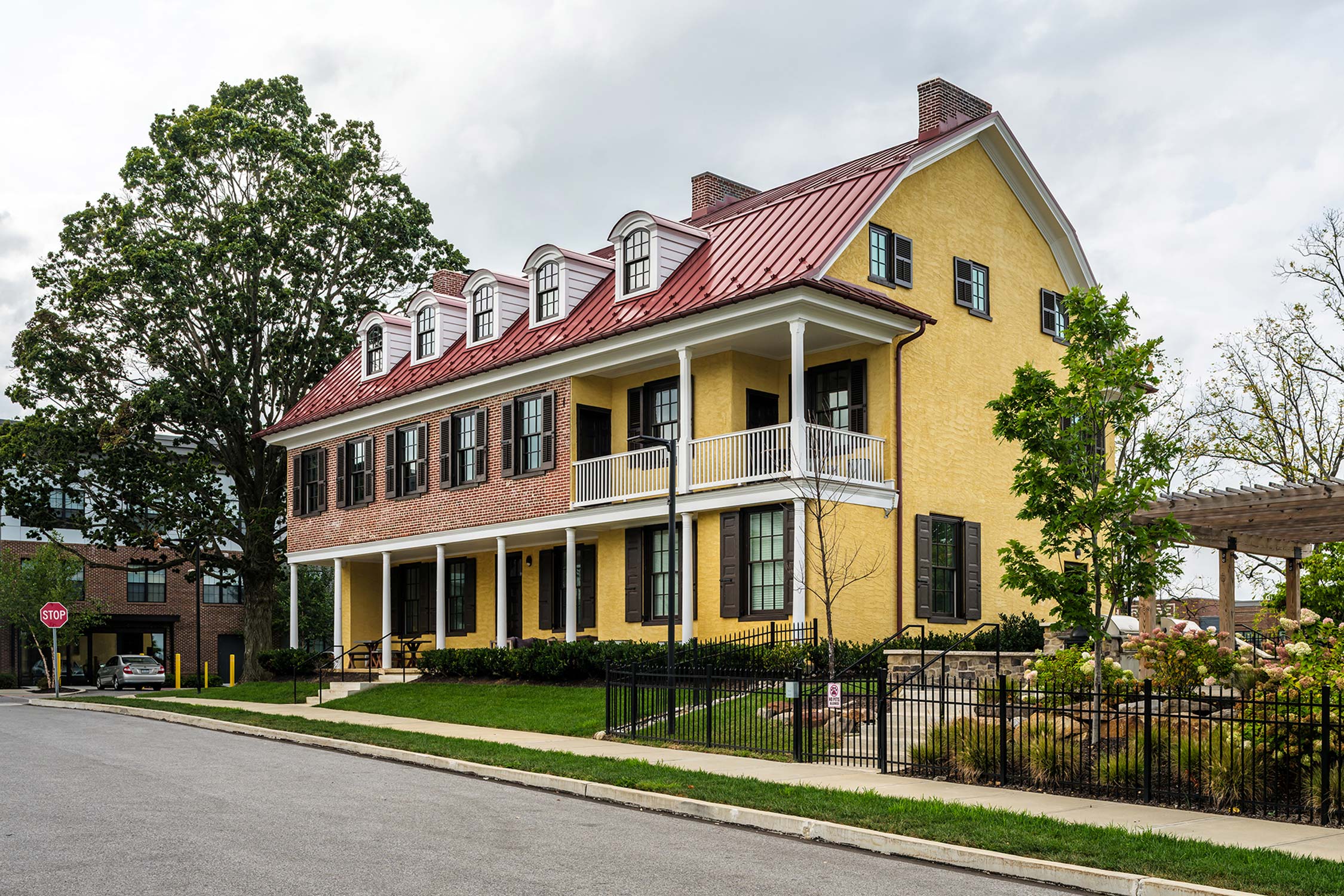 Yellow house with brick base, red roof, dormer windows, and balcony, near a street with a stop sign and trees.