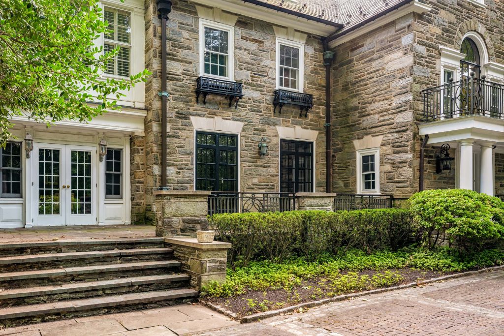 Stone exterior of the Augustinian Novitiate at Villanova University with double doors, steps, and large windows. A balcony, shrubs, and trees are visible.