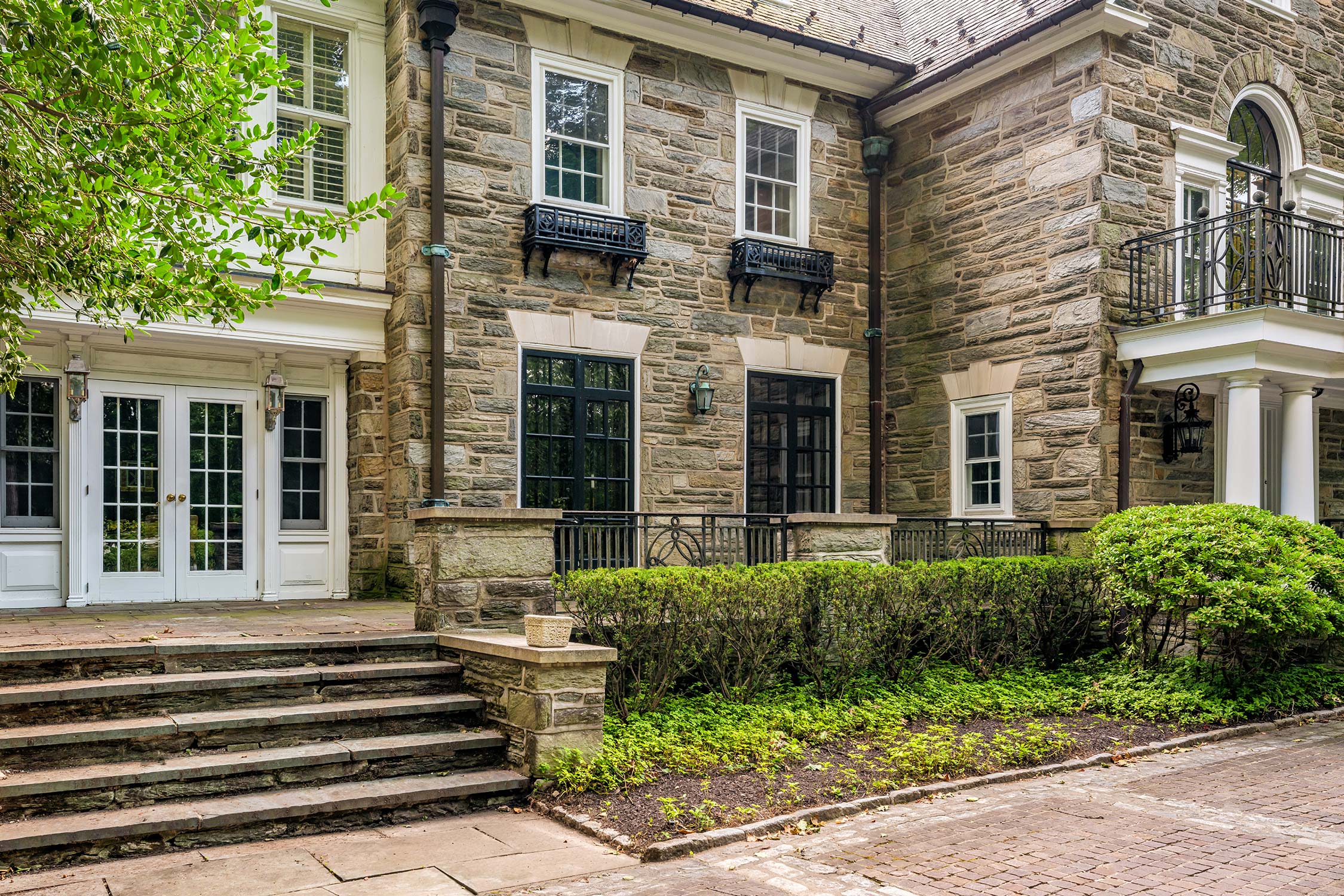 Stone exterior of the Augustinian Novitiate at Villanova University with double doors, steps, and large windows. A balcony, shrubs, and trees are visible.
