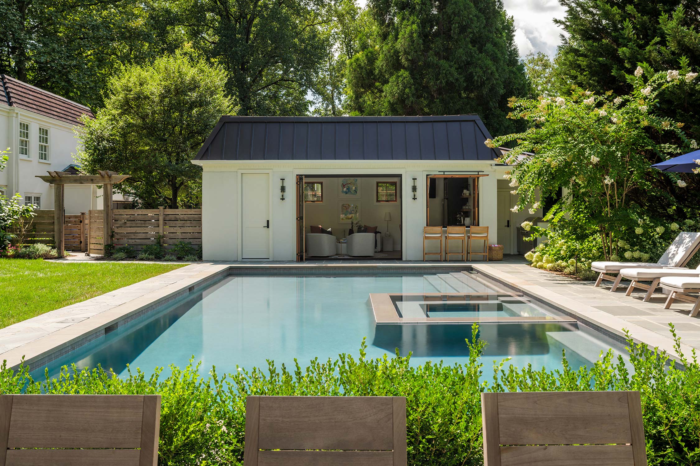 A poolside area with a white cabana, black roof, open seating, hot tub, and lush landscaping.