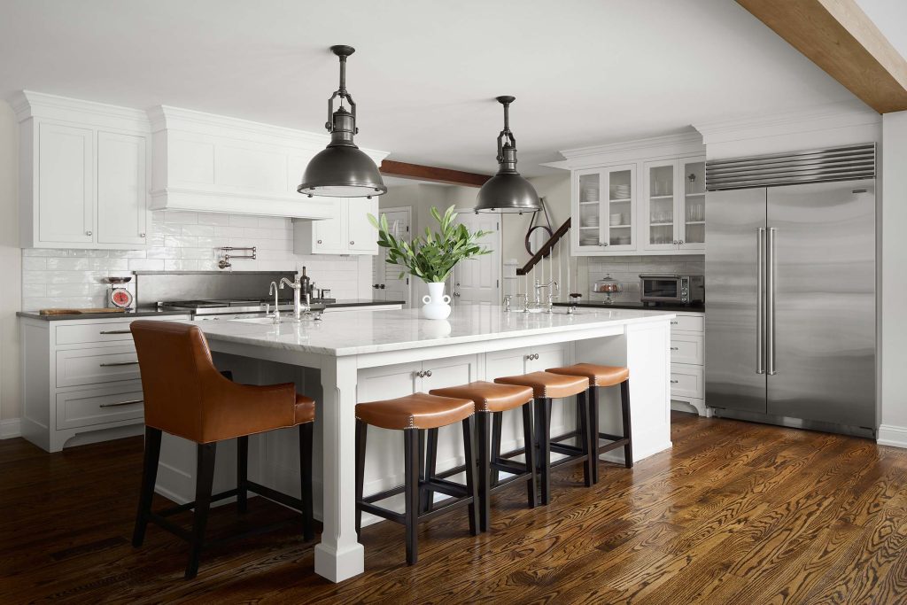Modern kitchen with white cabinets, large island, leather stools, pendant lights, and stainless steel appliances.