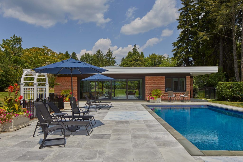 A poolside patio with lounge chairs, blue umbrellas, and a brick pool house with large windows. Flowers and greenery surround the area under a partly cloudy sky.