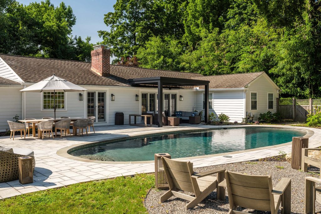 Backyard with a pool, white house, and pergola. Outdoor dining set with umbrella and wicker seating area on a stone patio, surrounded by trees.