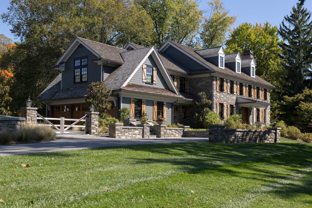 Large two-story house with gray siding, stone base, wooden shutters, and dormer windows, surrounded by lush greenery and a wooden gate.