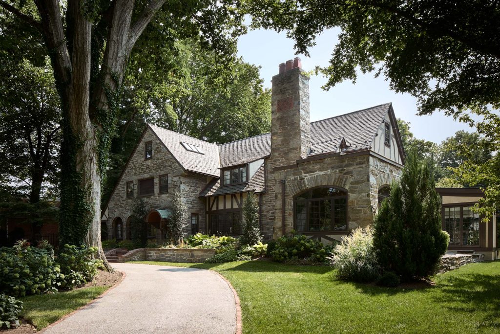 Stone house with gabled roof, chimney, ivy-covered tree, and lush garden with a curved driveway.