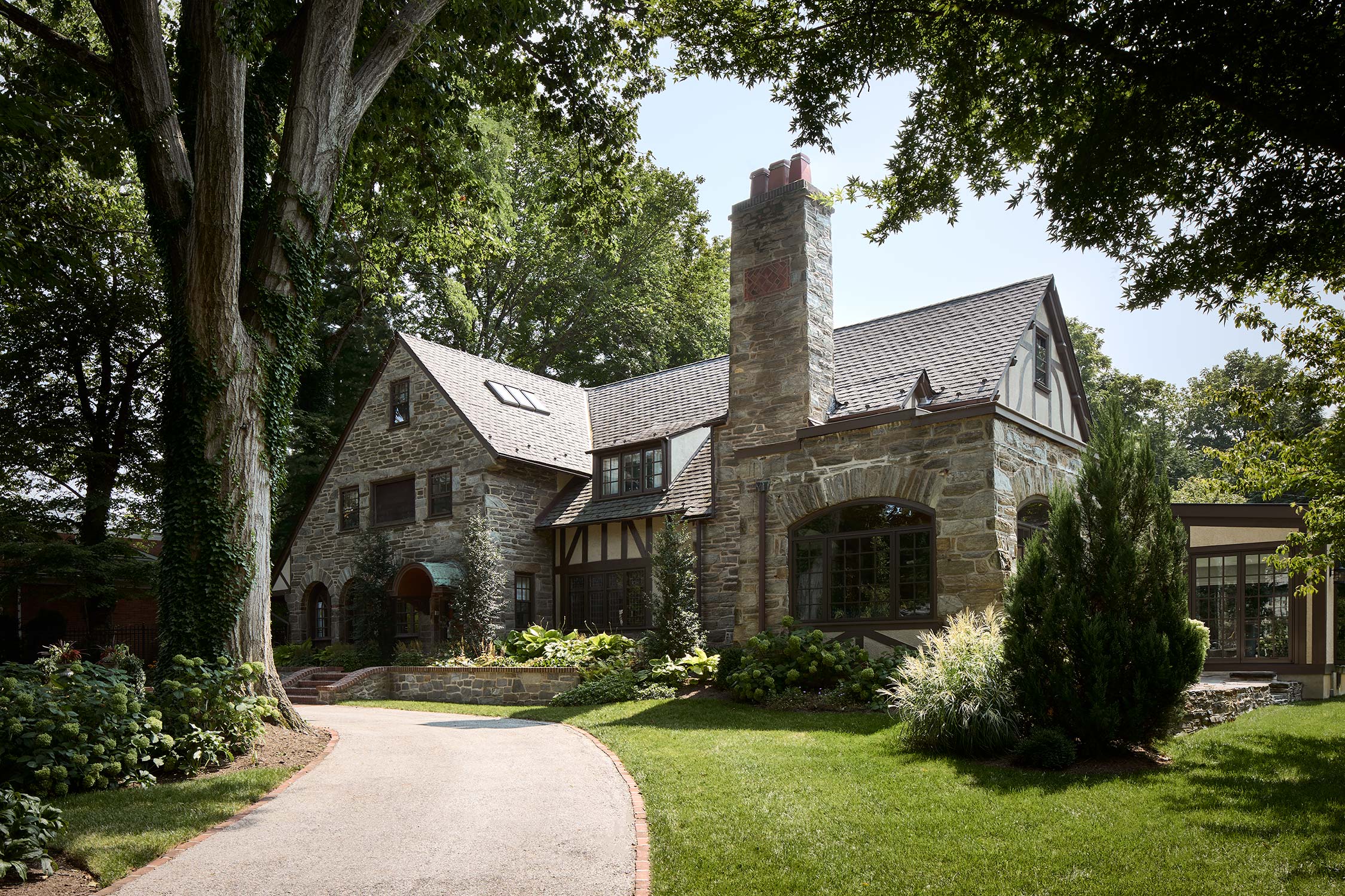 Stone house with gabled roof, chimney, ivy-covered tree, and lush garden with a curved driveway.