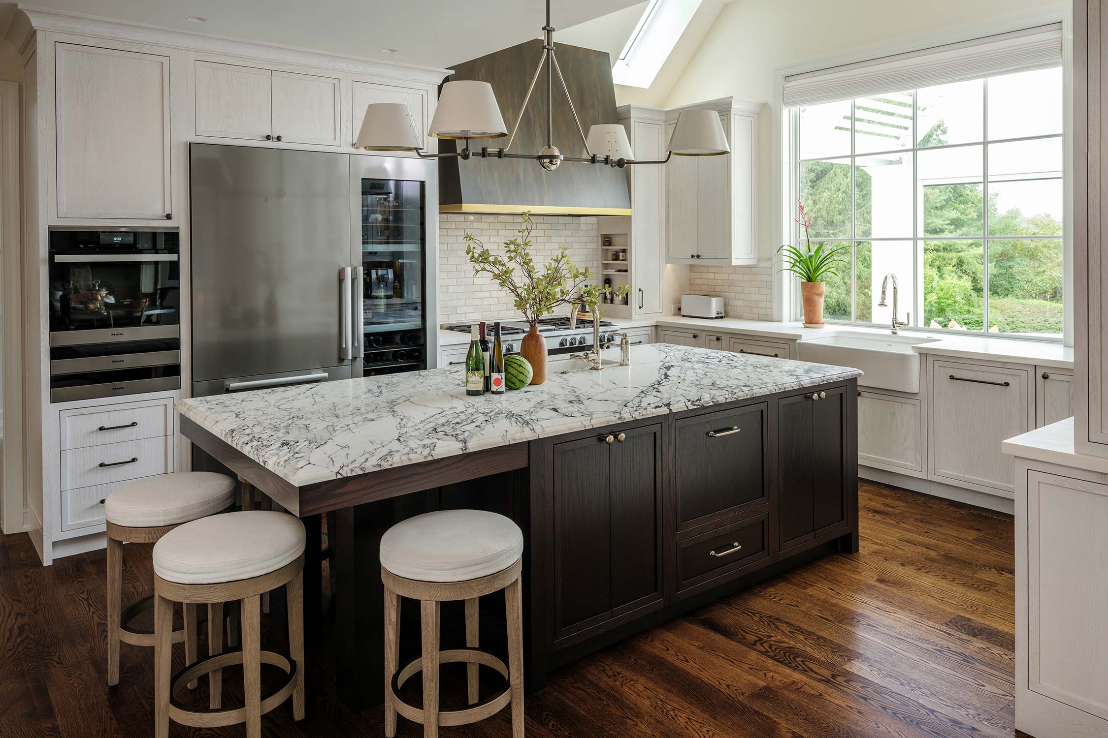 Modern kitchen with marble island, cushioned stools, stainless steel appliances, and large windows with greenery view.