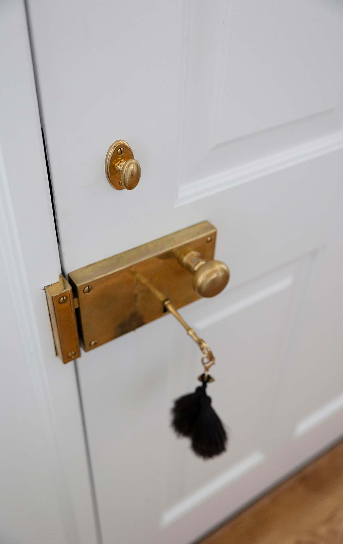 Close-up of a white door with an elegant brass handle and lock, featuring a black tassel keychain, set against wooden flooring.