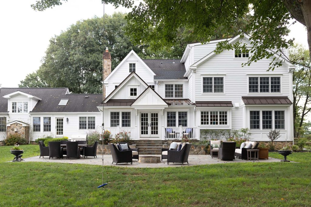 Spacious white house with stone accents, multiple windows, and a patio with wicker furniture and fire pit on a lush lawn.