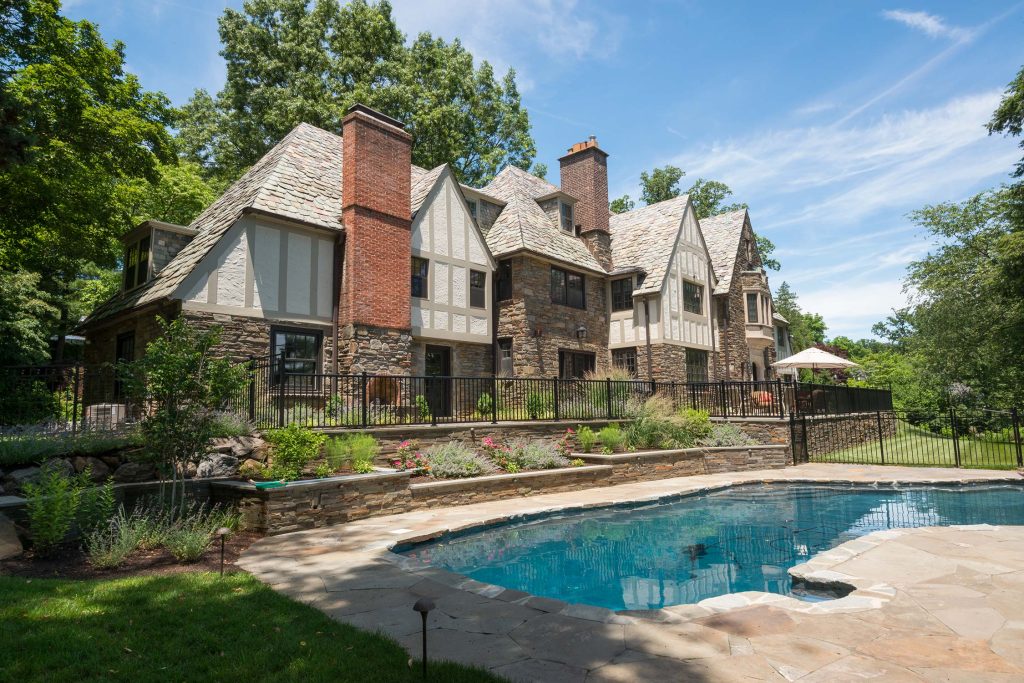 Elegant stone mansion with slate roof, brick chimneys, and lush gardens, featuring a sparkling pool under a clear blue sky.