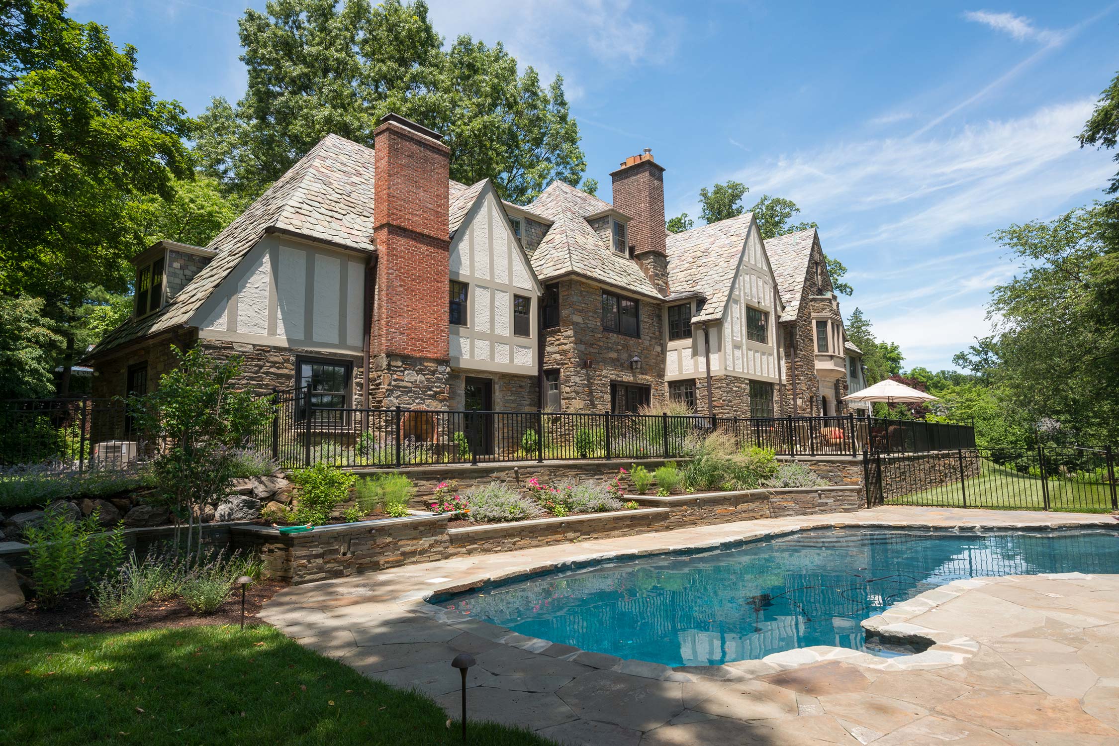 Elegant stone mansion with slate roof, brick chimneys, and lush gardens, featuring a sparkling pool under a clear blue sky.