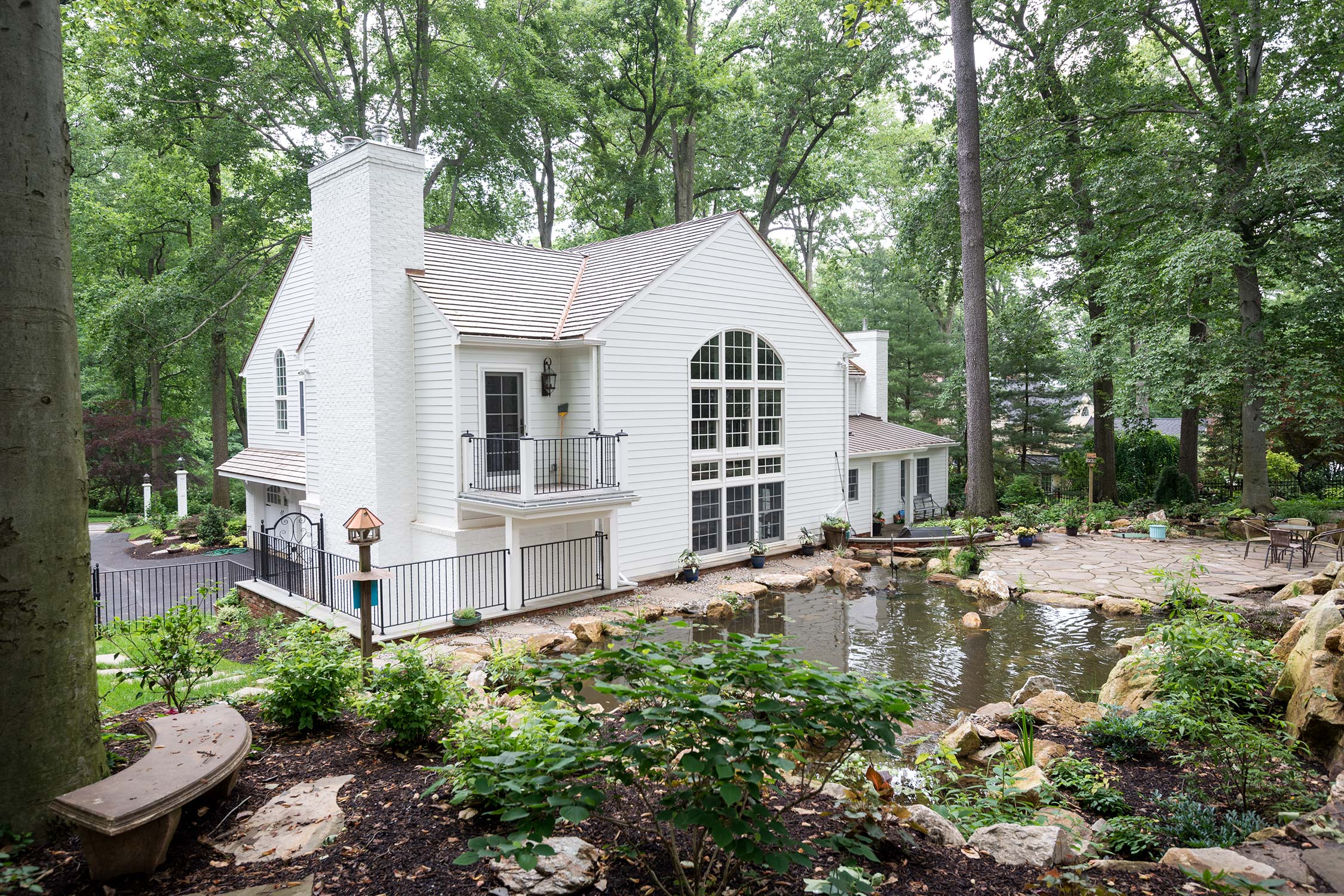 Elegant white house with large windows, balcony, and pond, surrounded by trees and lush landscaping.