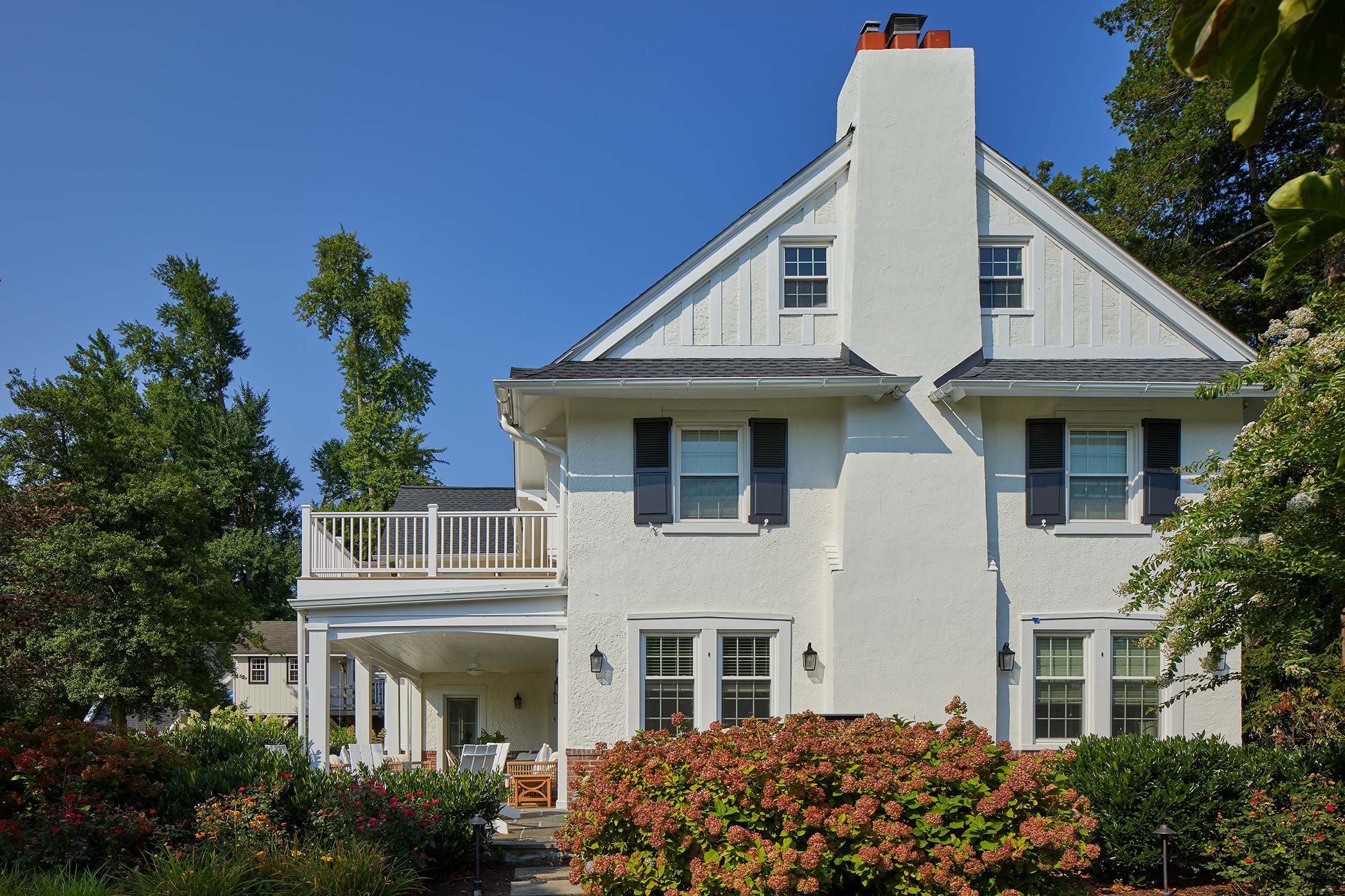 Yellow two-story house with wraparound porch, balcony, and dark roof, surrounded by green lawn, shrubs, and trees under a clear sky.