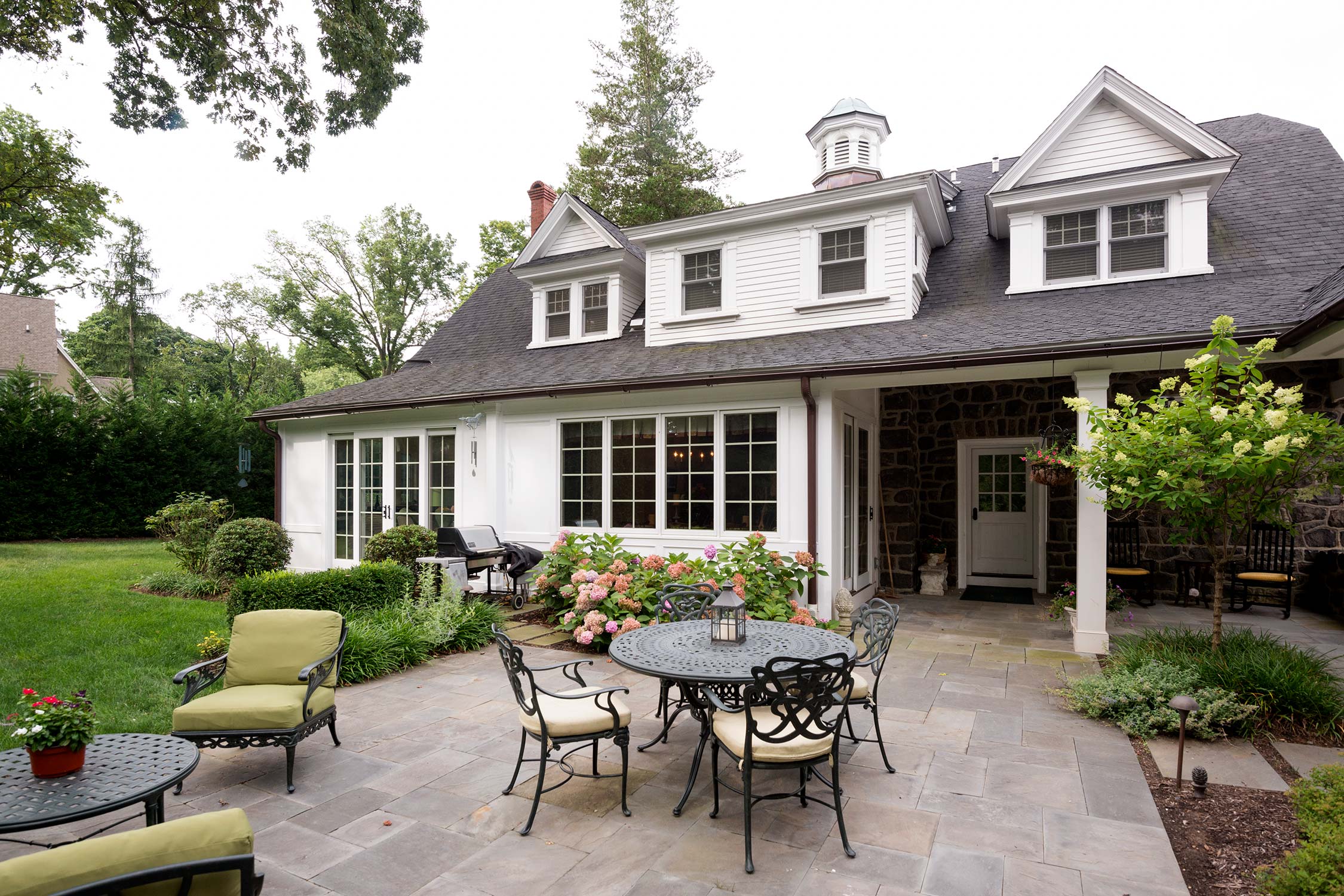 A stone and white house with a stone patio, outdoor table, chairs, and green cushions, surrounded by flowers and trees.