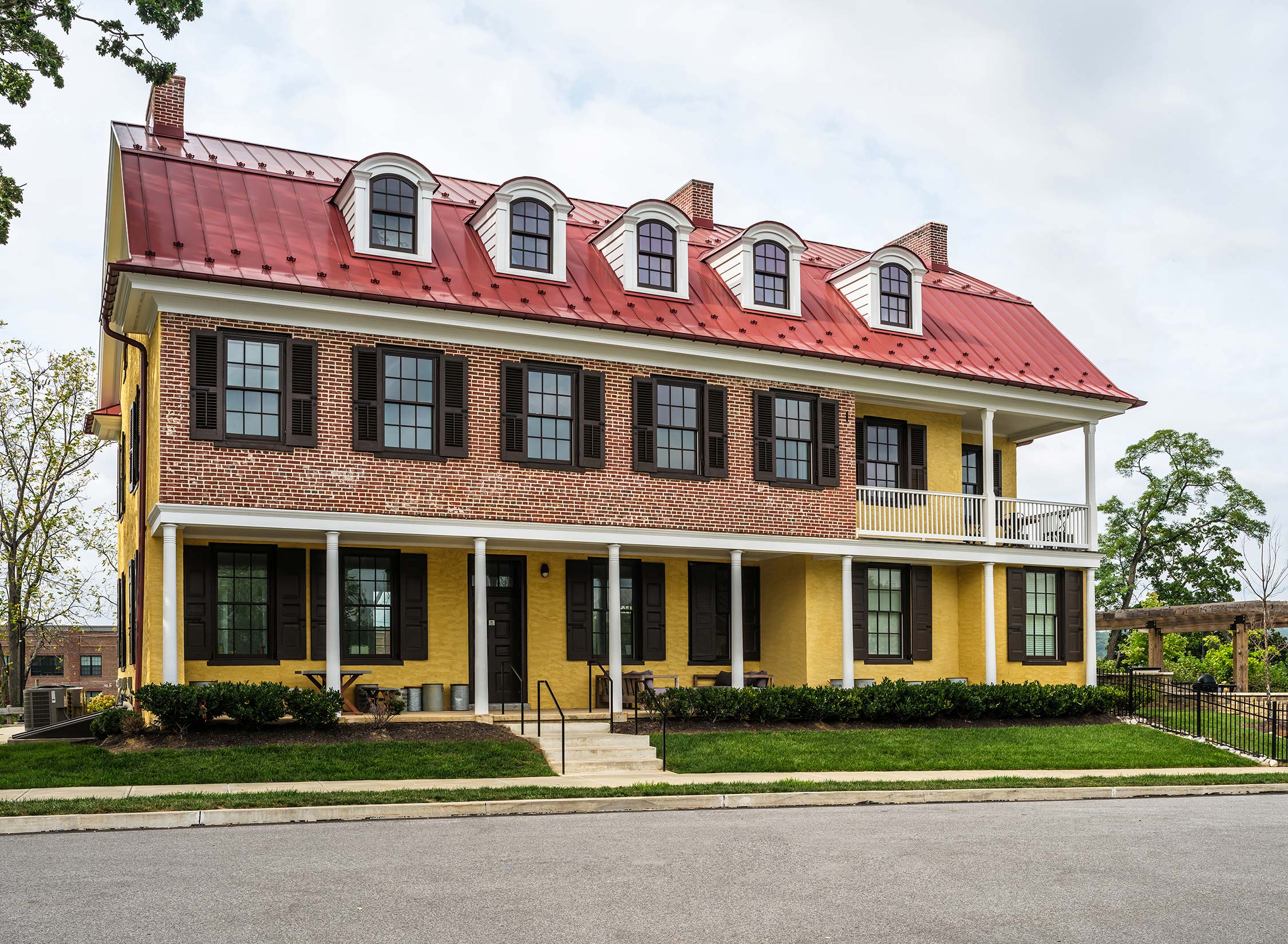 Yellow house with brick base, red roof, dormer windows, and balcony, near a street with trees.