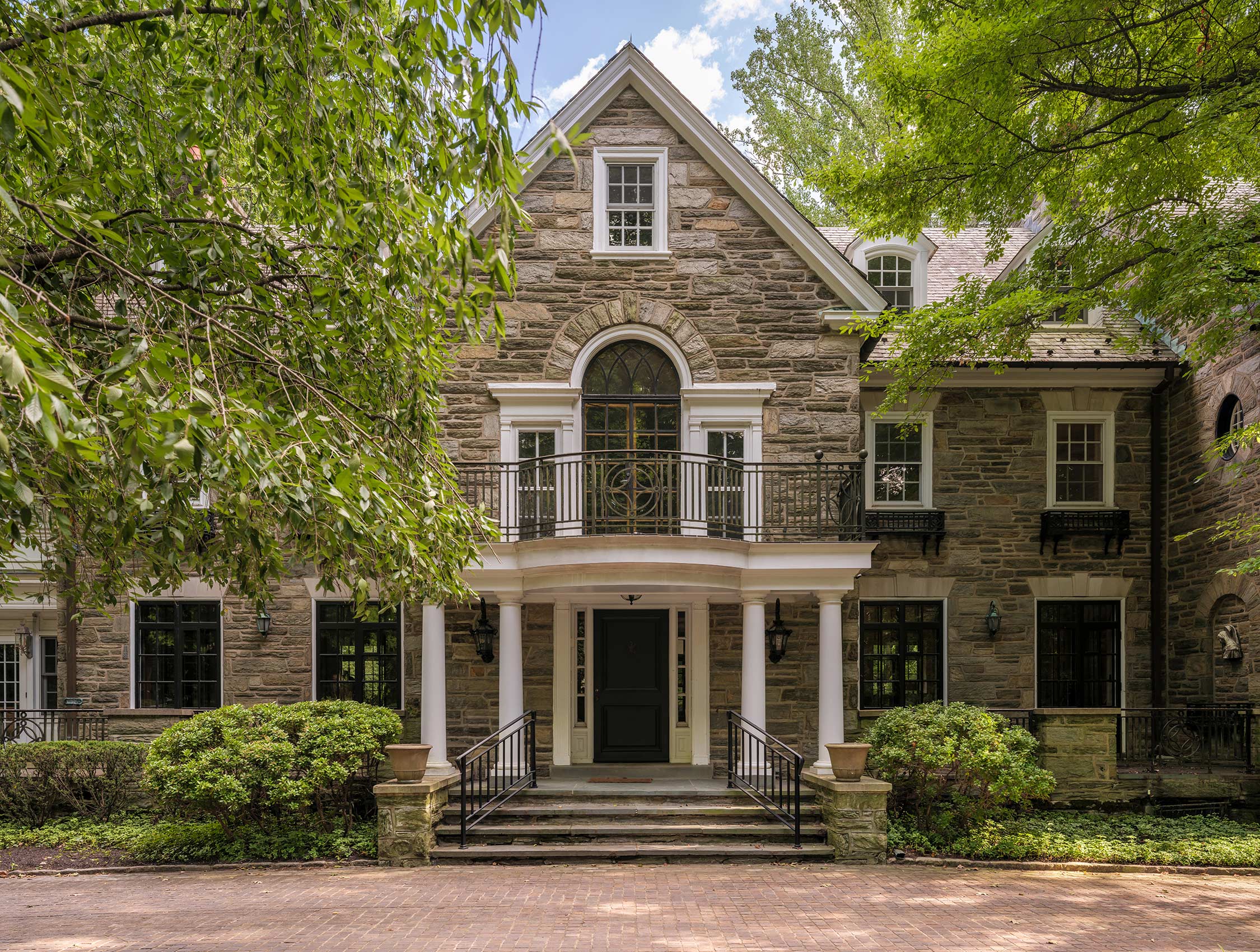 Stone facade of a building with a gabled roof, arched window, and balcony. The entrance has columns, steps, and a black door, surrounded by shrubs and trees.