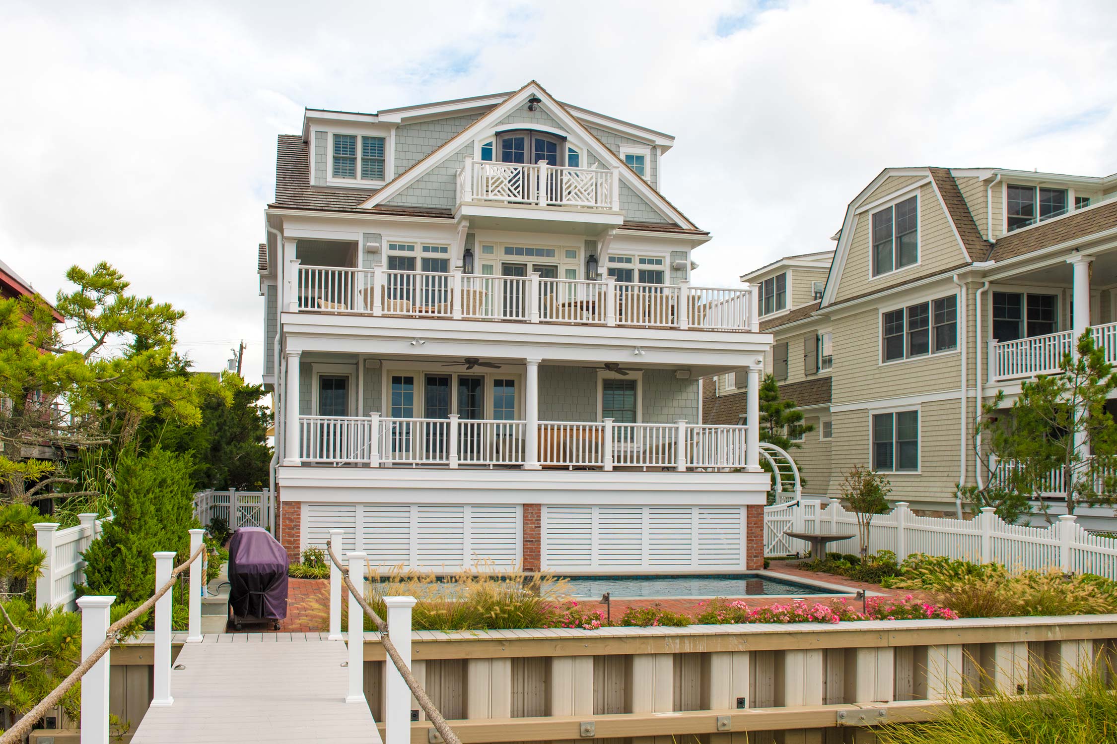 Multi-story beach house with gray shingles, white railings, and balconies. Small pool and garden in the backyard, wooden walkway to a covered grill.