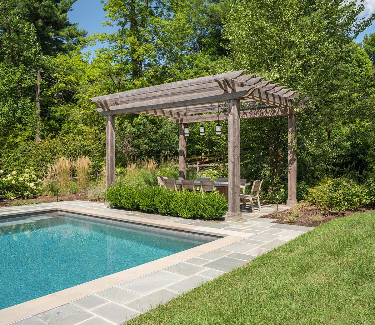 A poolside pergola with wooden beams, pendant lights, a dining table, chairs, and lush greenery.