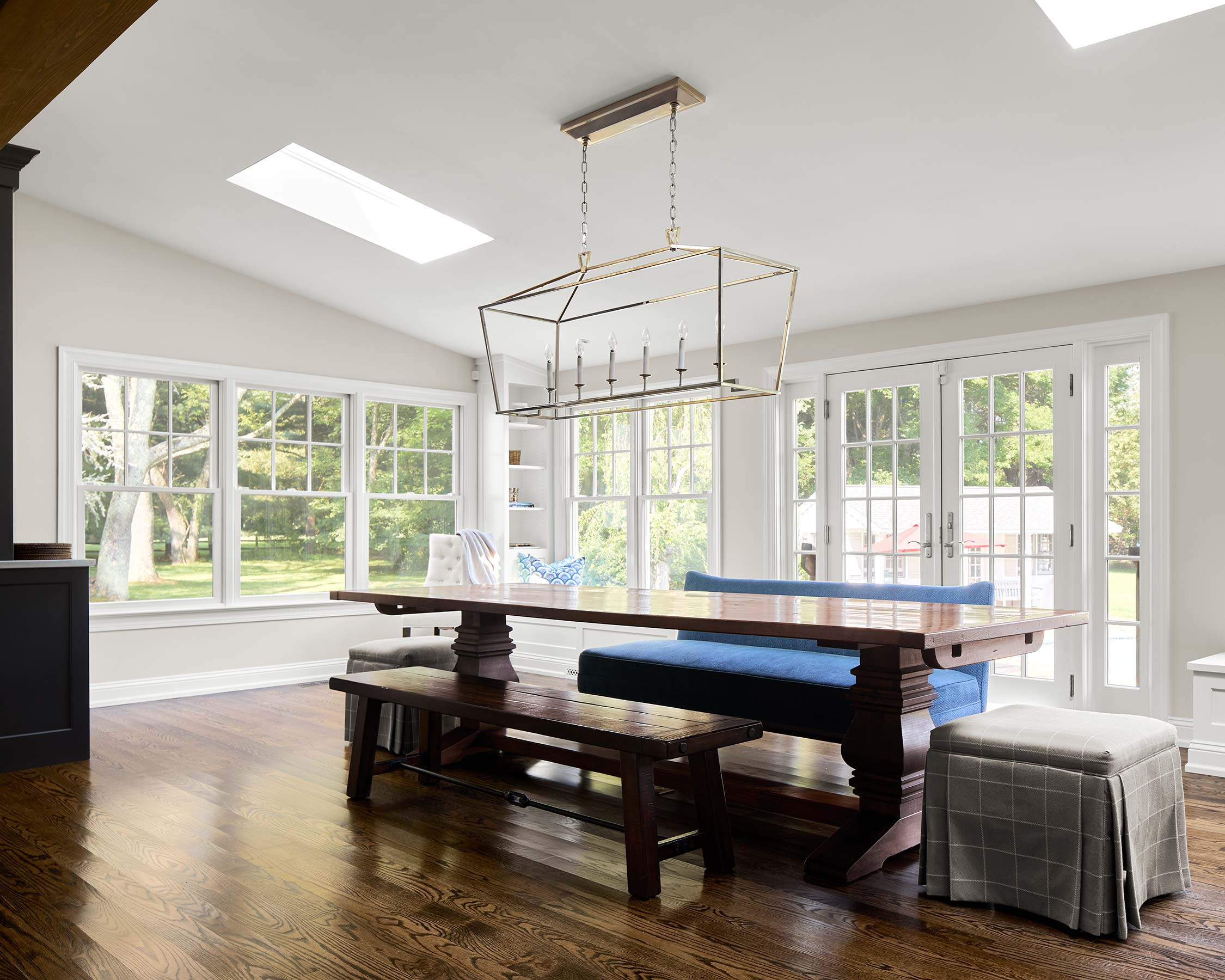 A bright dining room with large windows, a wooden table, blue cushioned bench, wooden benches, a gray ottoman, and a modern chandelier.