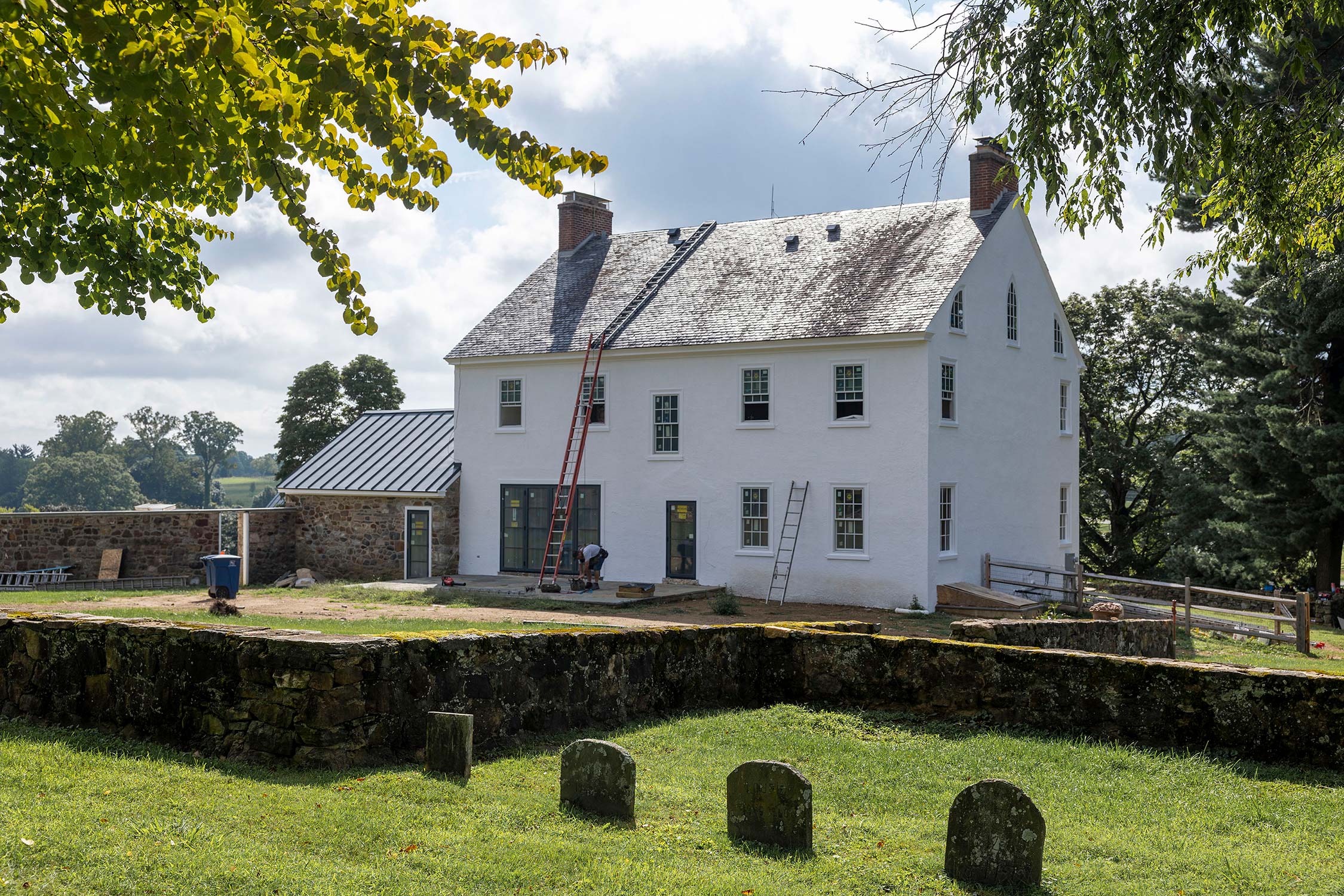 Worker with ladders at a white historic home with stone facade and metal roof, surrounded by stone walls and greenery.