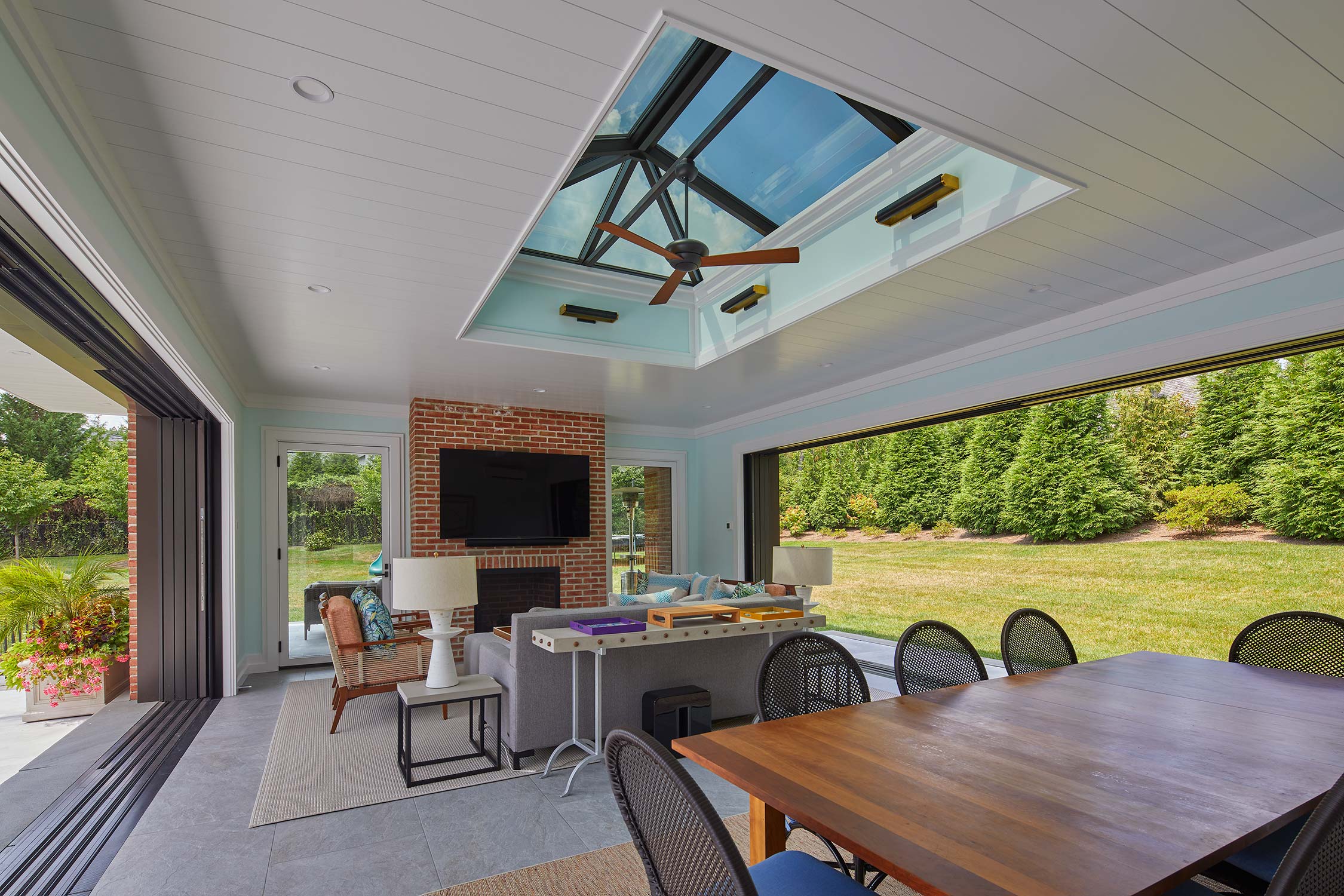Indoor-outdoor room with skylight, brick fireplace, TV, dining table, and chairs, opening to a grassy lawn with trees.