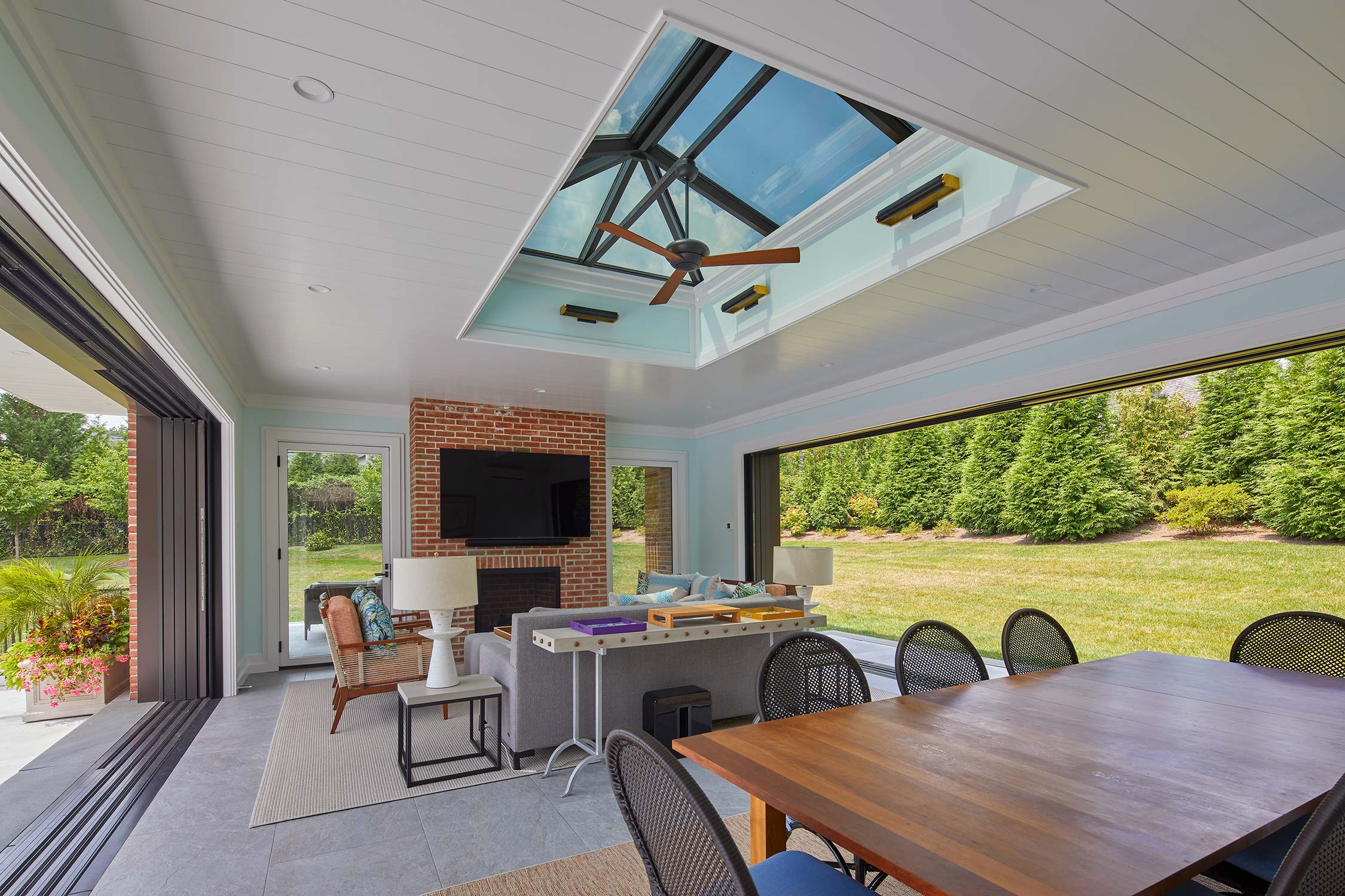 Indoor-outdoor room with skylight, brick fireplace, TV, dining table, and chairs, opening to a grassy lawn with trees.