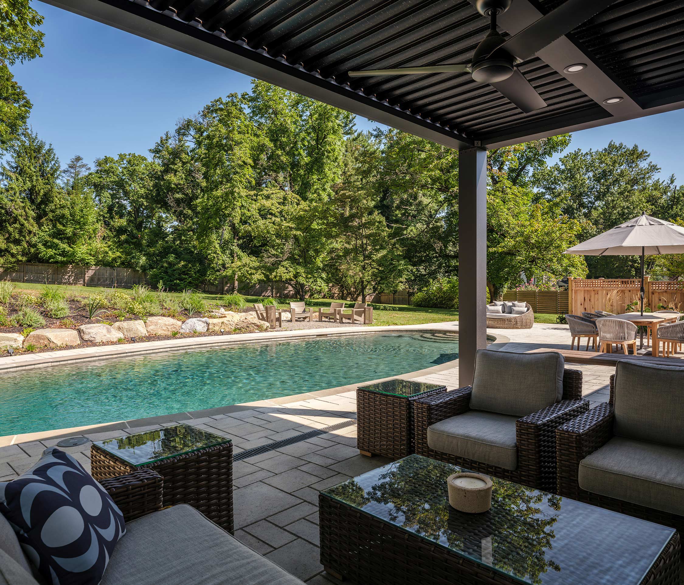 Outdoor patio with wicker furniture, glass tables, and a pool. A pergola with a ceiling fan overlooks a lush backyard with trees and a dining area.