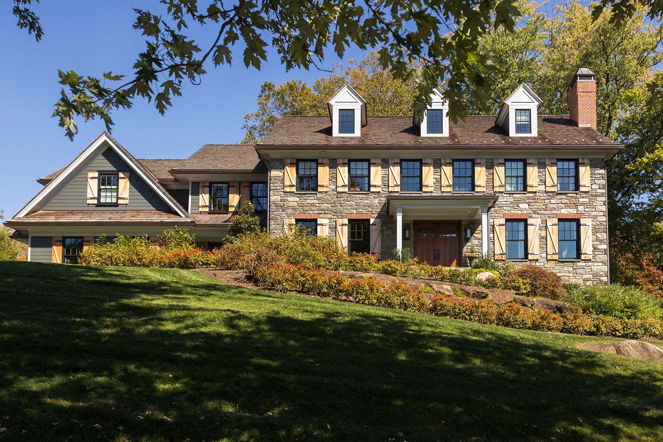 Large two-story house with gray siding, stone base, wooden shutters, dormer windows, and surrounded by lush greenery.