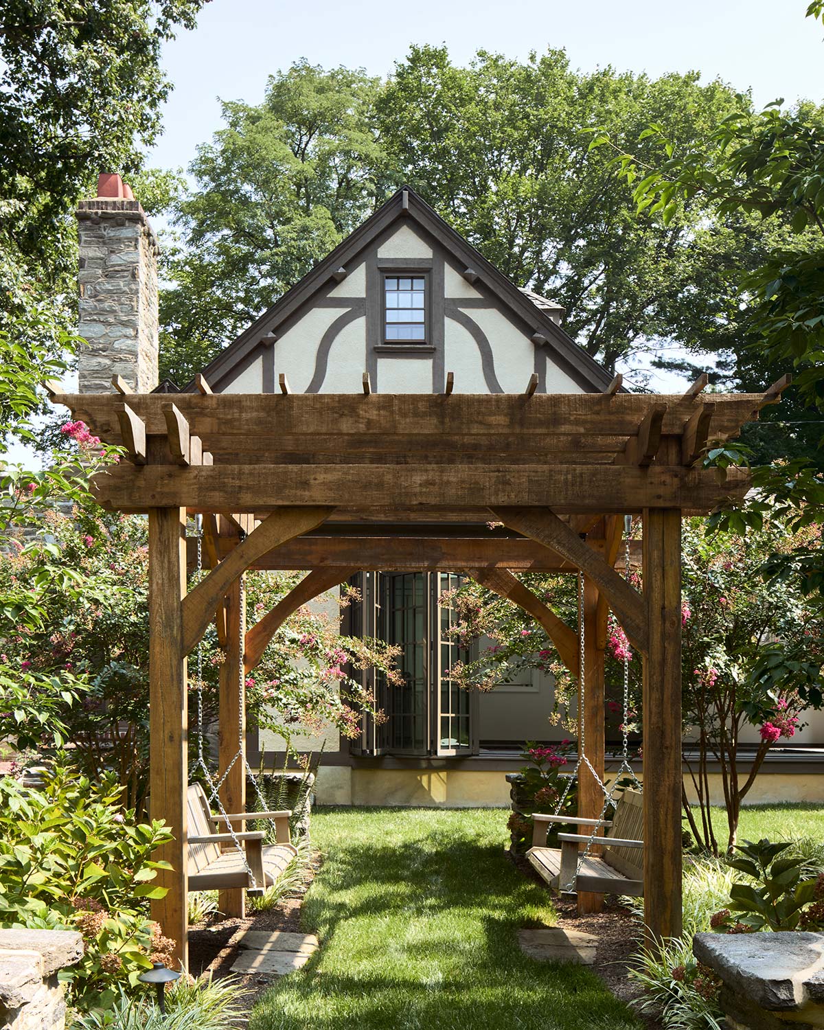 Tudor-style house with wooden pergola, two hanging swings, green lawn, and blooming shrubs.