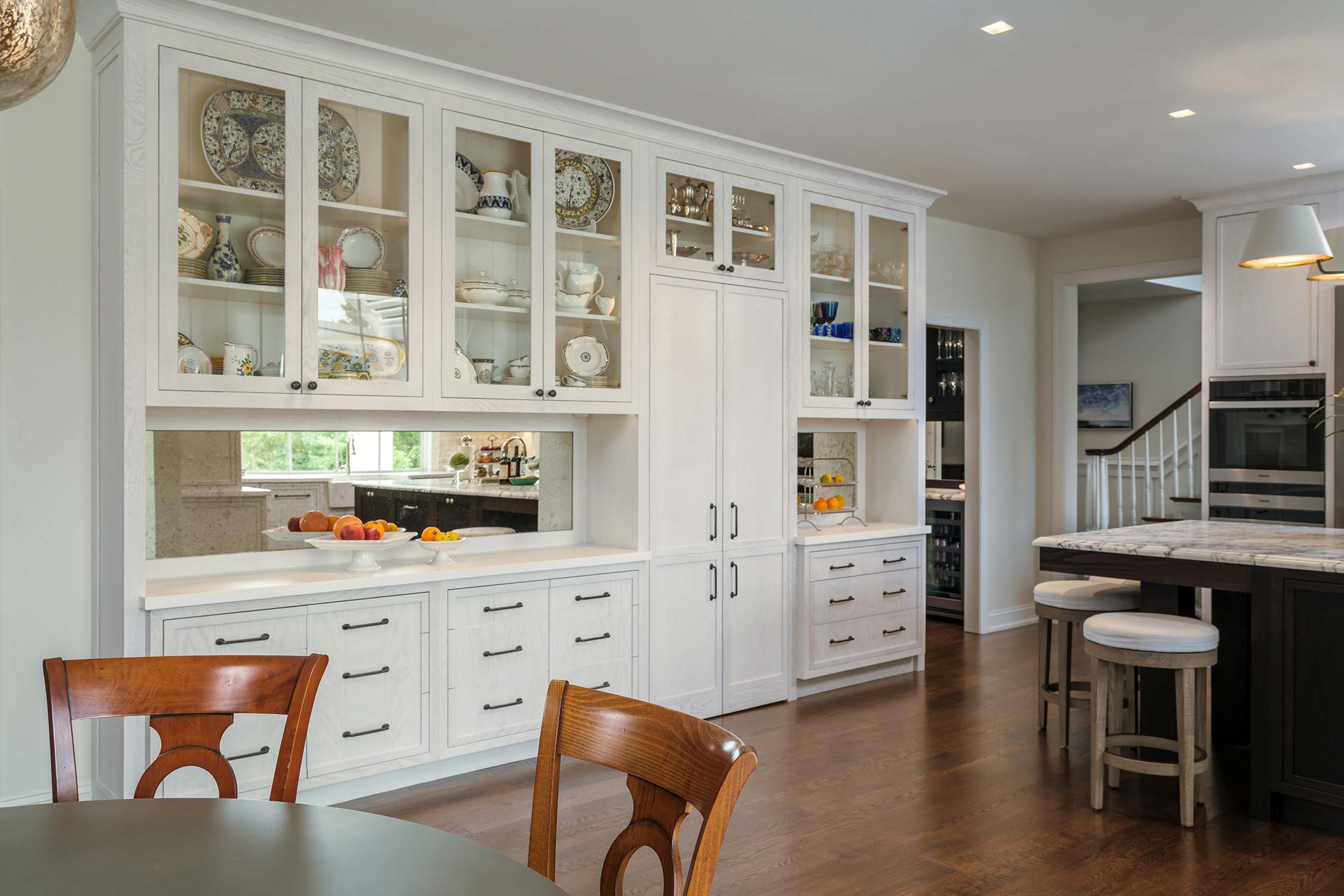 Elegant kitchen with white cabinets, glass shelves displaying dishes, a counter with fruit, and cushioned stools at a dark island.