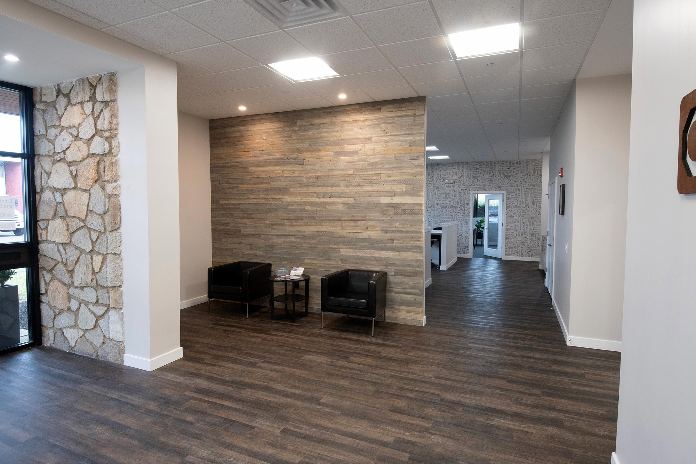 Office lobby with wooden floor, stone wall, two black chairs, and a small table.