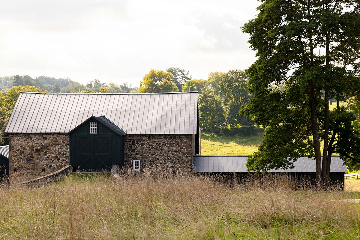 Rustic stone barn with a metal roof and black doors, set against a backdrop of tall grass, trees, and rolling hills under a cloudy sky.