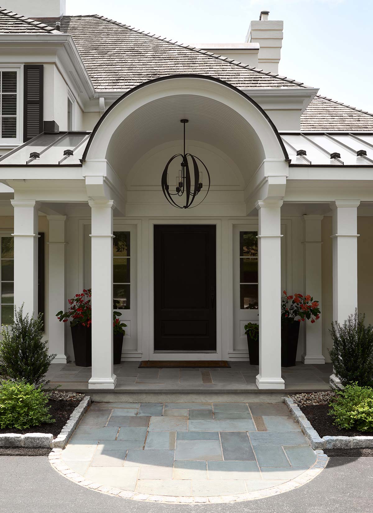 A house entrance with an arched porch, white columns, black door, potted plants, and stone pathway.