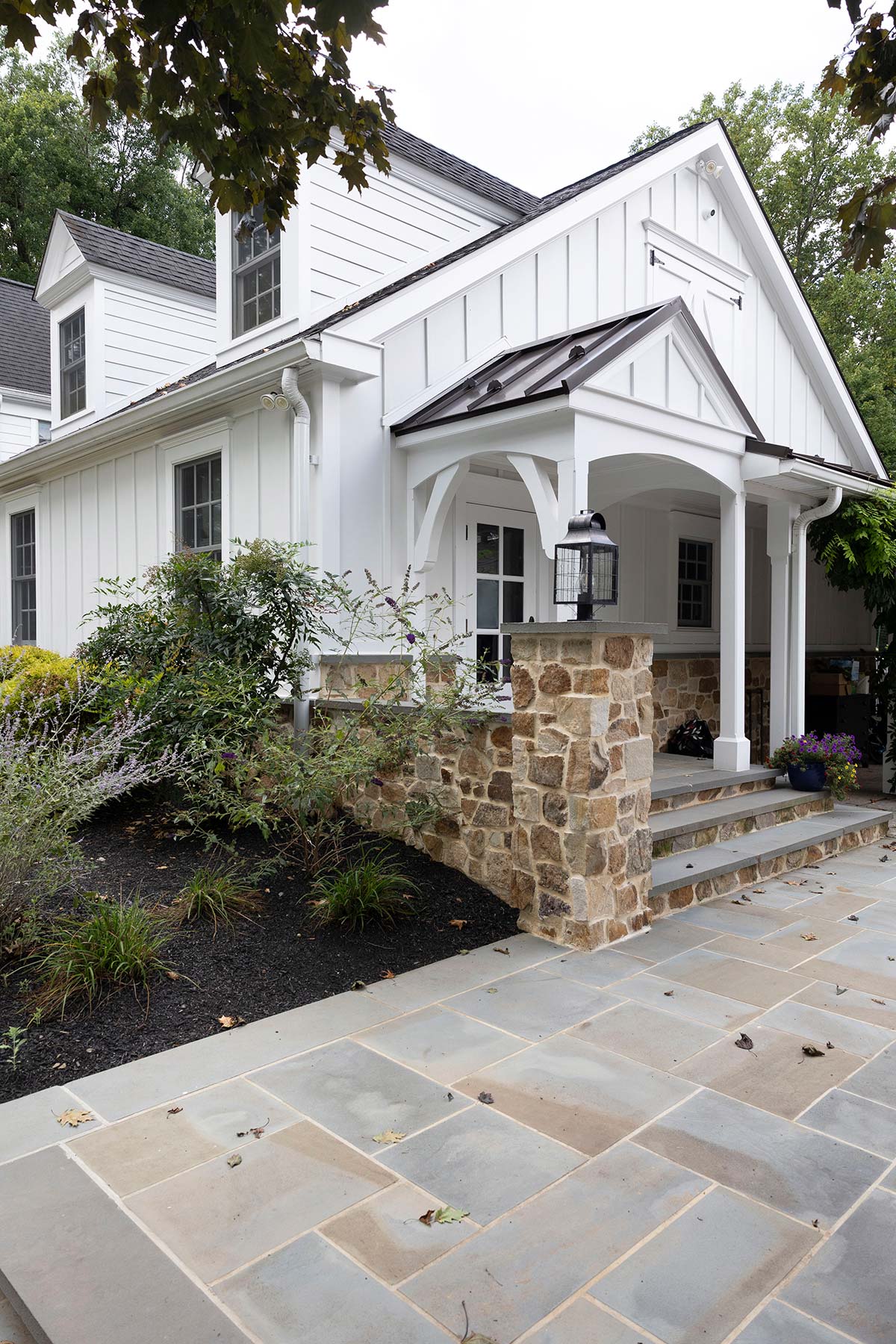 White house with stone base, dormer windows, covered porch with lantern, and a stone-paved walkway surrounded by greenery.