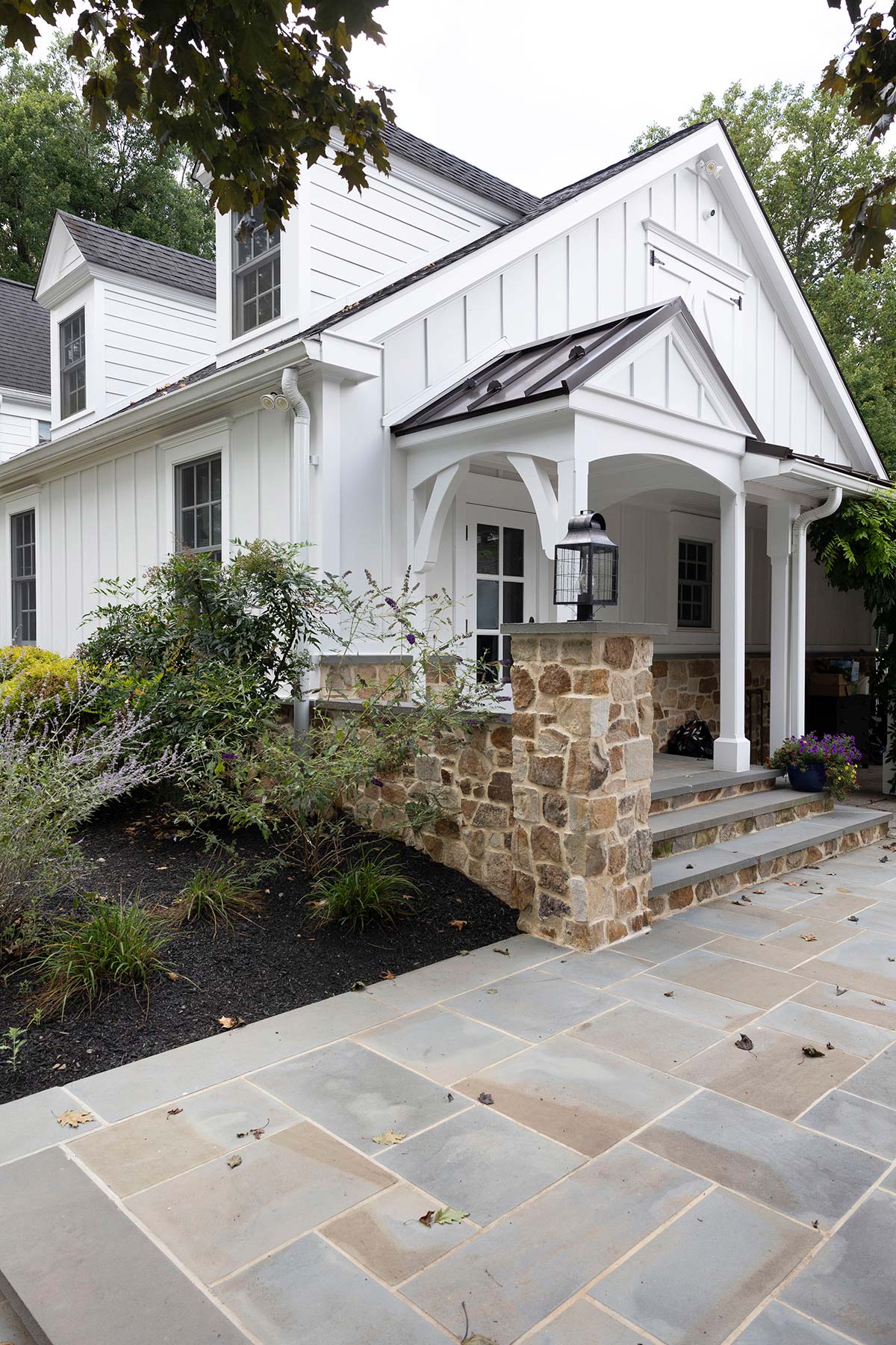 White house with stone base, dormer windows, covered porch with lantern, and a stone-paved walkway surrounded by greenery.