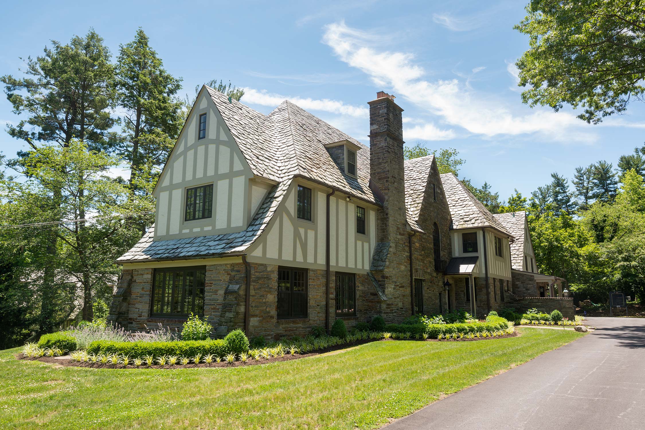 Charming stone and timber home with slate roof and brick chimney, surrounded by lush greenery and manicured gardens under a bright sky.