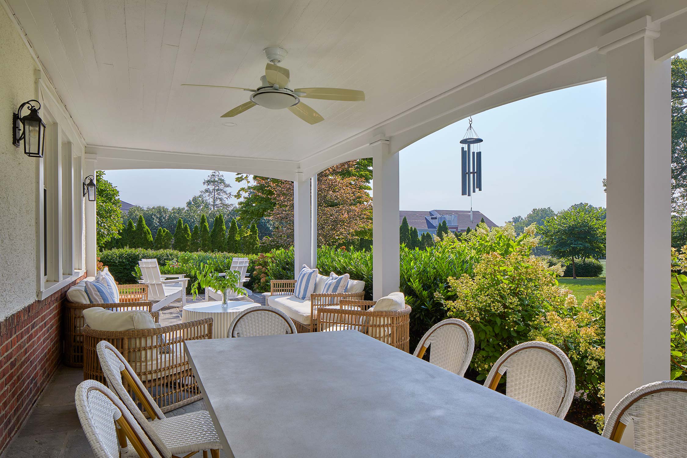 A covered porch with a ceiling fan, wicker furniture, a long dining table, and a view of a lush green backyard with shrubs and trees.