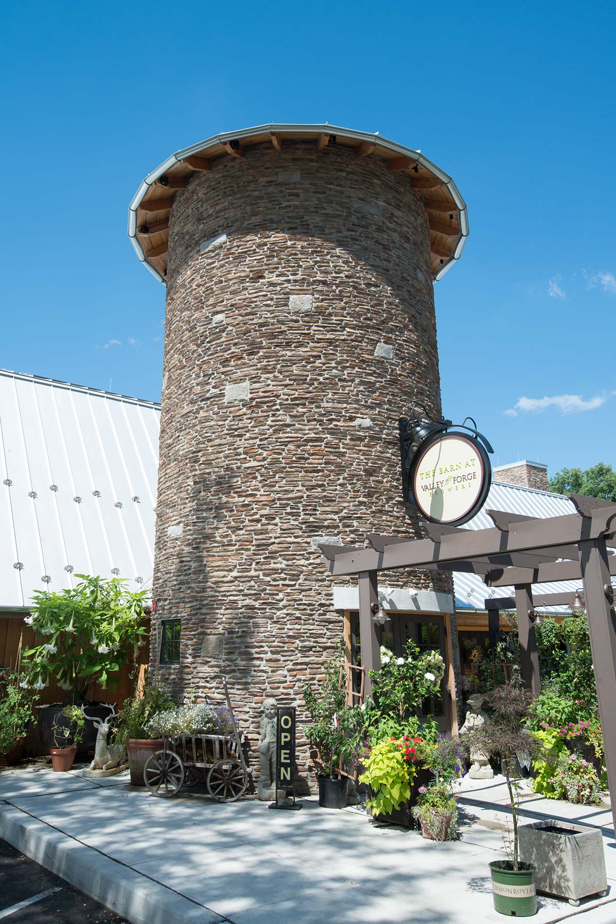 Stone silo at Valley Forge Flowers with a sign, potted plants, and a wooden cart under a pergola on a sunny day.