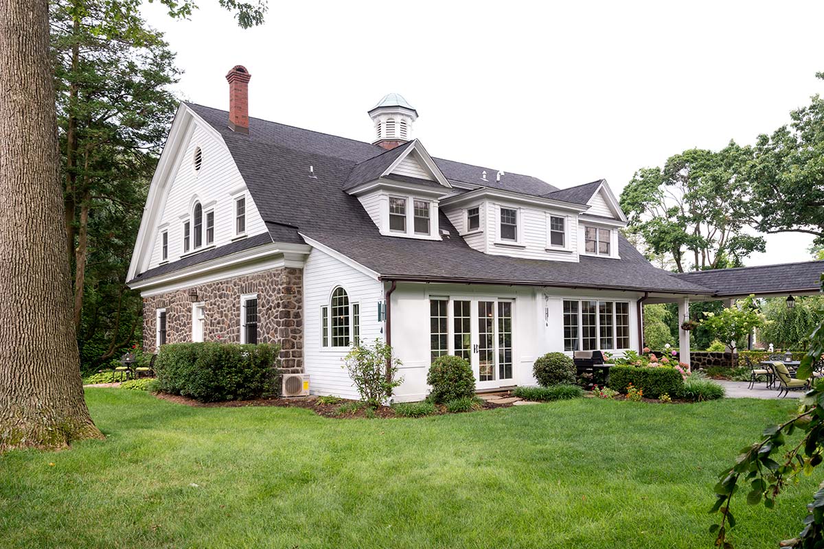 A stone and white house with a large lawn, patio, and outdoor seating, surrounded by trees and shrubs.
