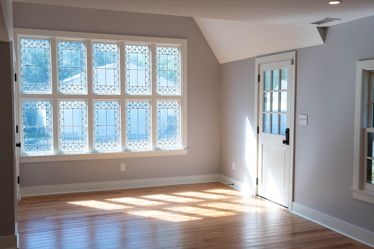 Sunny cottage interior with hardwood floors, light gray walls, ornate multi-pane windows streaming sunlight, and a white door.