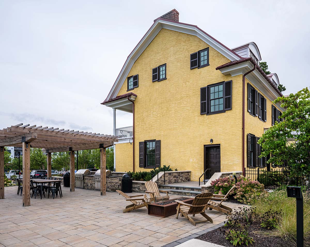 Yellow house with patio, wooden pergola, fire pit, chairs, dining table, and stone grill area, surrounded by plants.