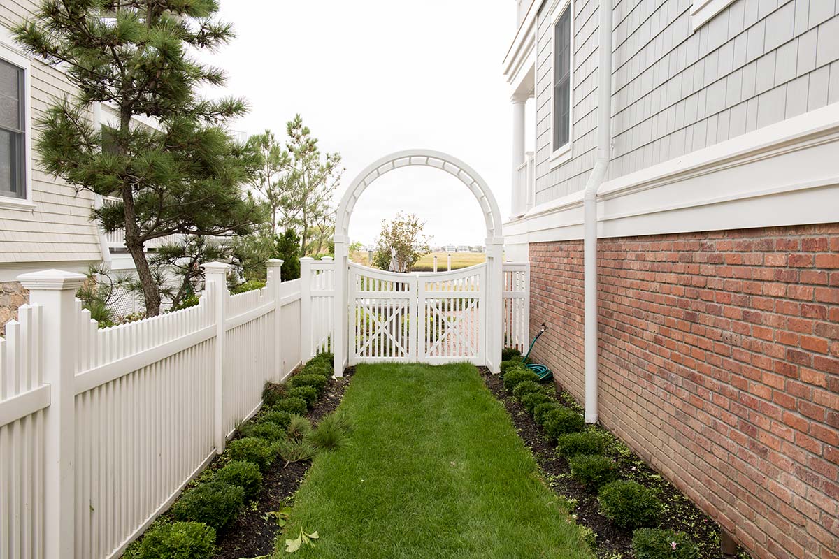 Narrow garden path with green lawn and shrubs, leading to a white arched gate between a white picket fence and a brick wall, flanked by gray shingle houses.