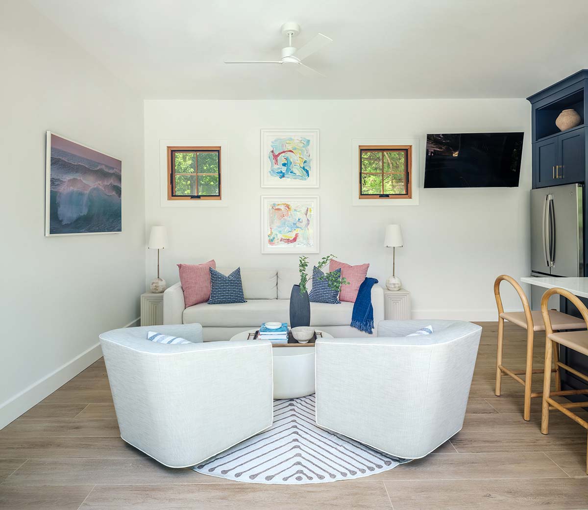 A cozy living room with two white armchairs, a sofa, pink and blue pillows, art on the walls, and a ceiling fan.