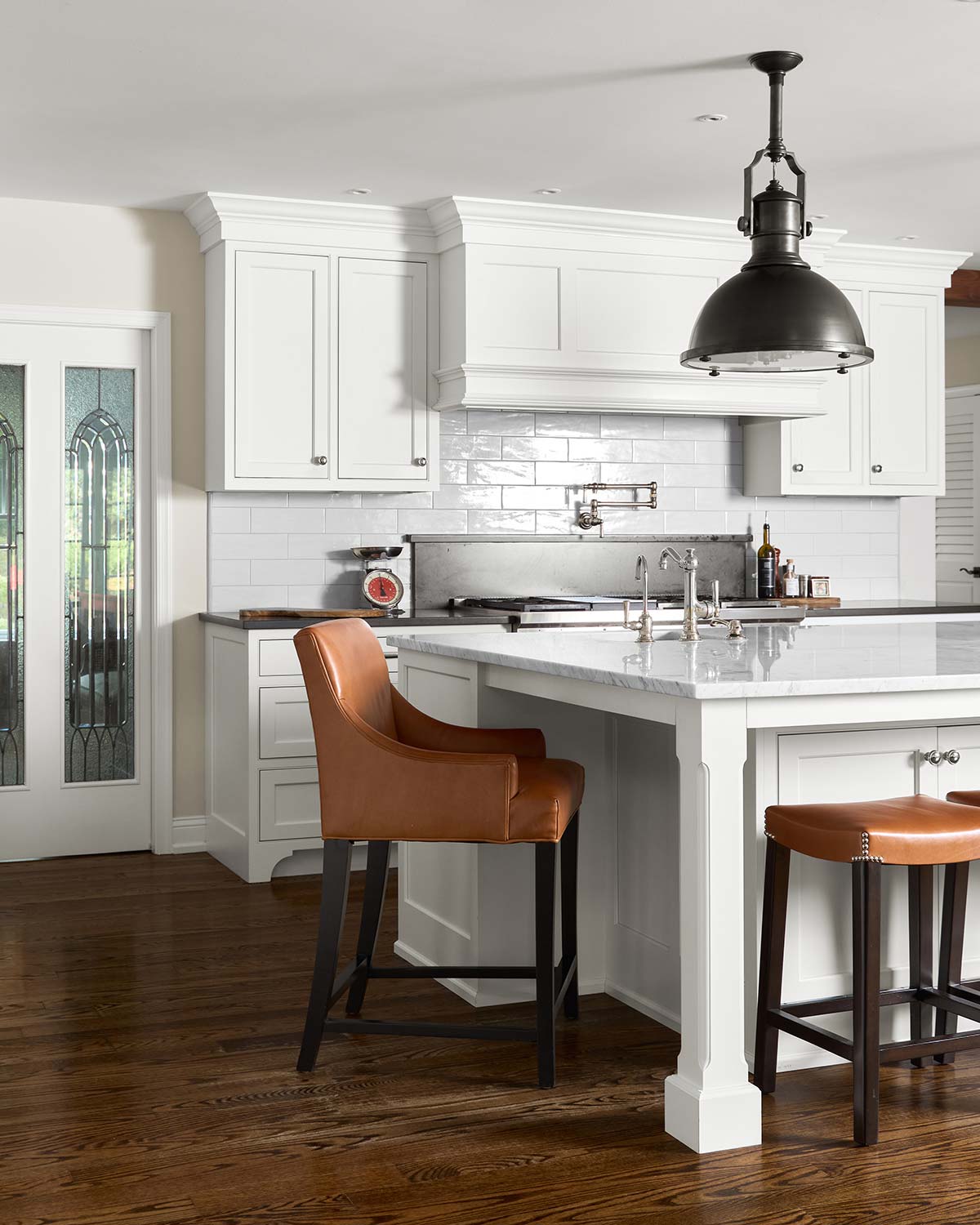 Modern kitchen with white cabinets, marble island, two brown leather bar stools, and a black pendant light.
