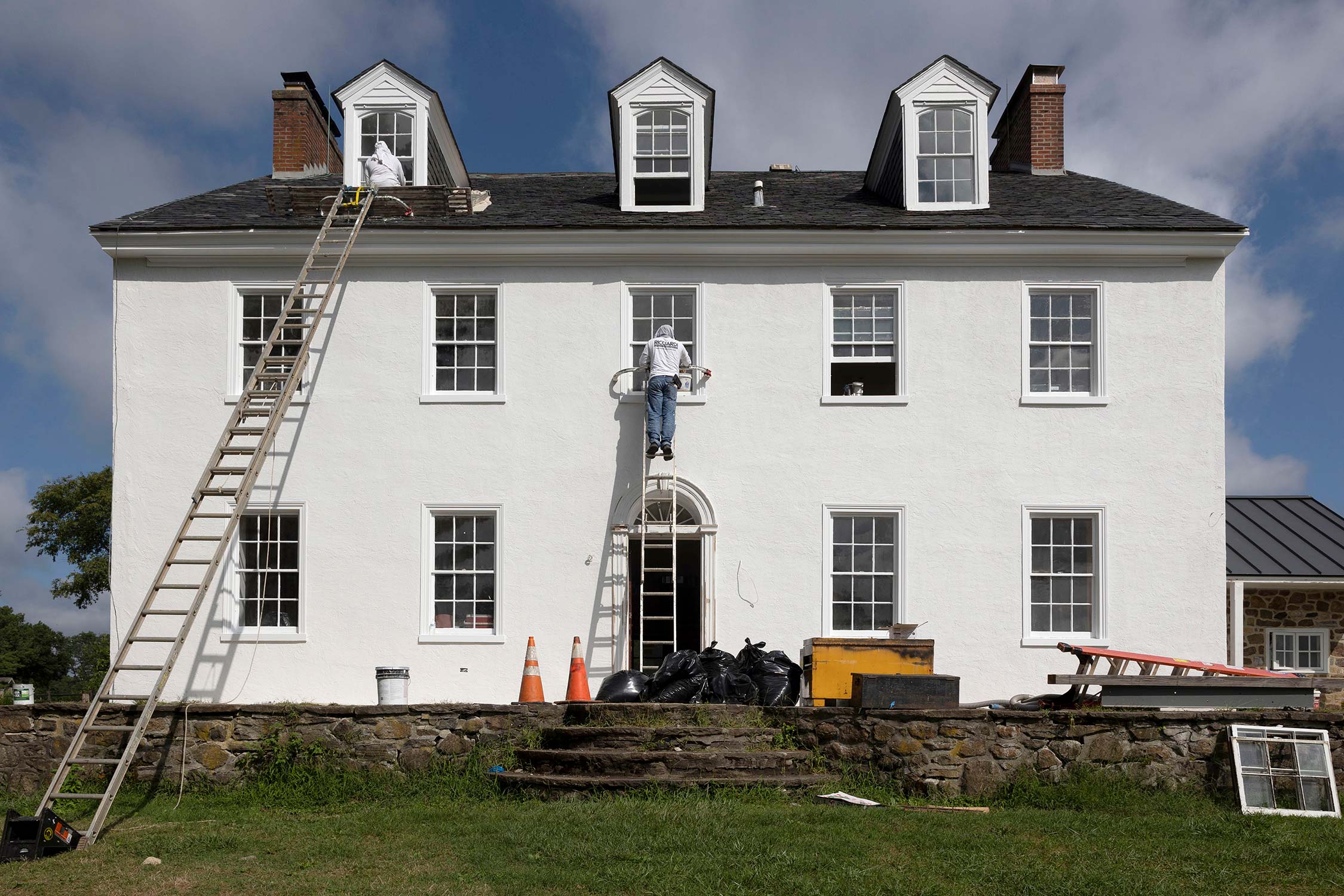 Two workers on ladders painting a white historic home with dormer windows, surrounded by stone wall and construction materials.