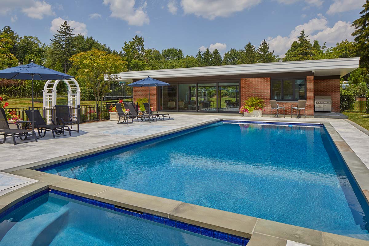 A poolside patio with lounge chairs, blue umbrellas, and a brick pool house with large windows. Flowers and greenery surround the area under a partly cloudy sky.