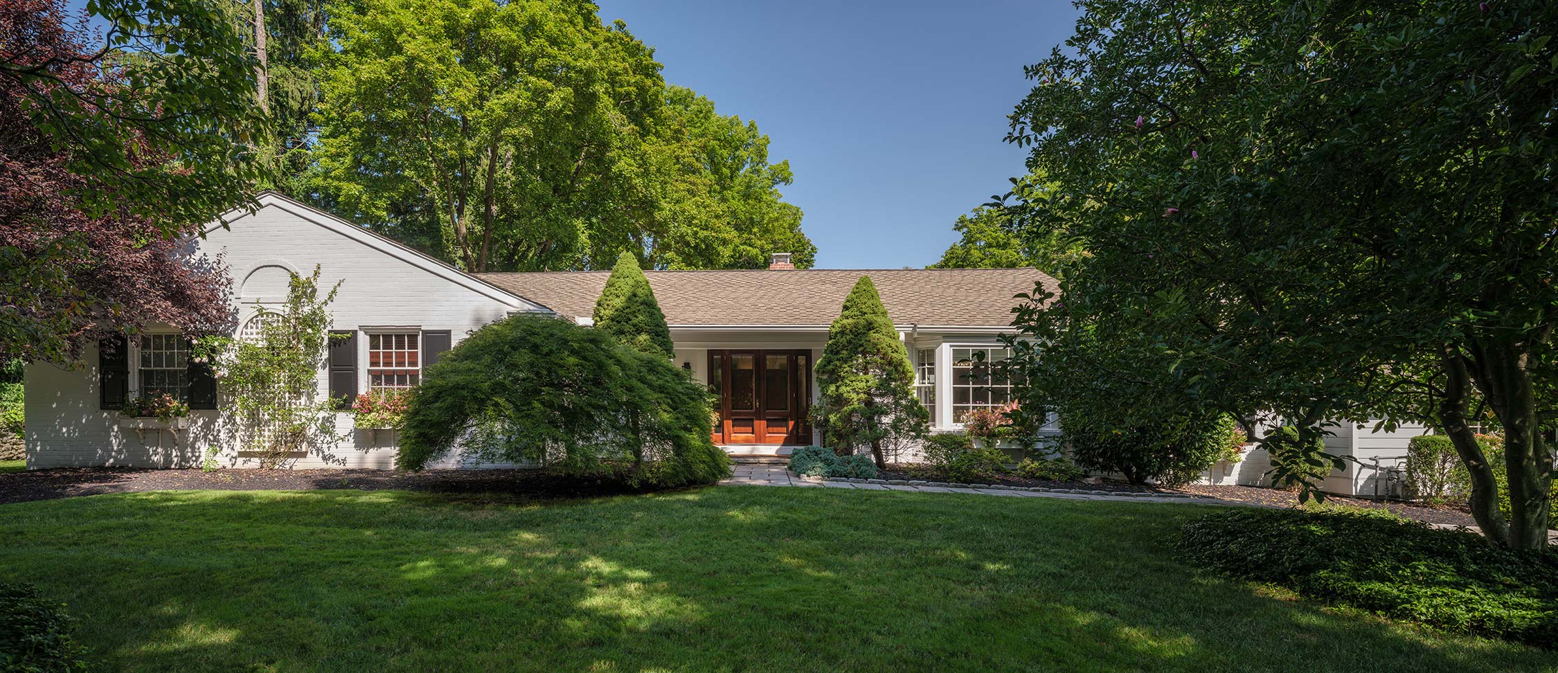 White mid-century house with wooden door, surrounded by lush green lawn, trees, and shrubs under a clear blue sky.
